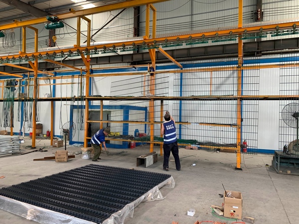 Workers hang 2D fence panels on conveyor racks before surface treatment.