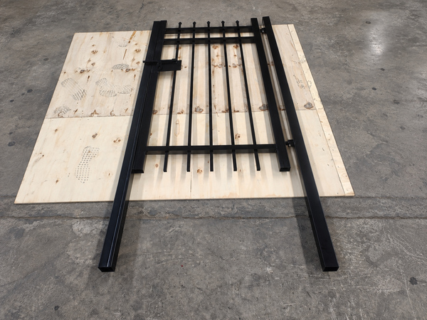 Powder coated black steel tubular fence gate displayed on plywood in workshop.